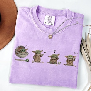 Puede incluir: Una camiseta lila Comfort Colors con un gr&aacute;fico de un personaje en varias poses. El personaje se muestra en una c&aacute;psula, jugando con una pelota, confundido y diciendo "coo!". Un collar plateado con un colgante est&aacute; sobre la camiseta.