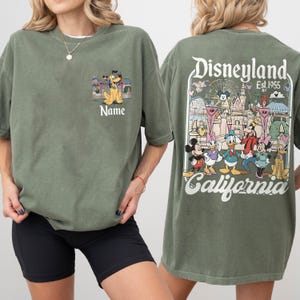 May include: Olive green t-shirt featuring a Disneyland graphic. The front has a cartoon image of Pluto and the word "Name". The back displays the Disneyland logo with various cartoon characters and the text "California".
