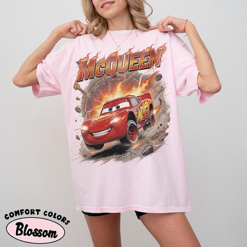Może przedstawiać: Jasnor&oacute;żowa koszulka z grafiką Zygzaka McQueena z filmu Auta. Projekt zawiera słowo "McQueen" w płomiennej czcionce nad samochodem. Koszulka ma r&oacute;wnież słowa "Comfort Colors" i "Blossom" wydrukowane na dole.
