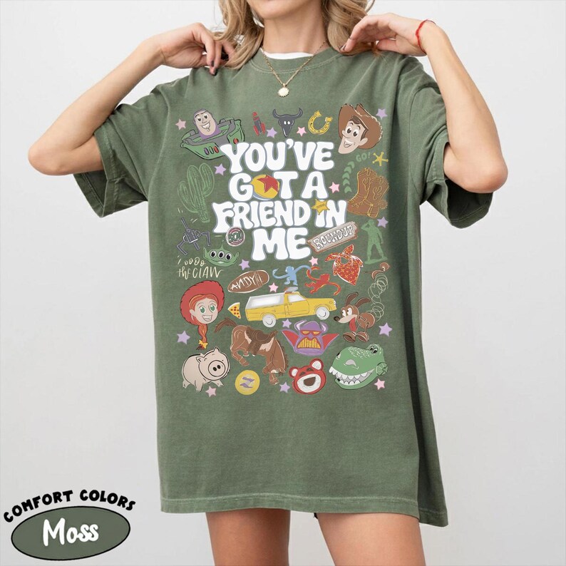 Op de afbeelding: Een olijfgroen T-shirt met korte mouwen en een grafische print met de tekst "YOU'VE GOT A FRIEND IN ME", omringd door diverse kleurrijke illustraties van stripfiguren, een gele vrachtwagen, een cactus en sterren in een collage-stijl.