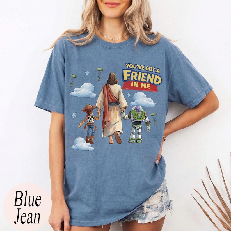 May include: Blue jean coloured t-shirt with a graphic that says "YOU'VE GOT A FRIEND IN ME". The graphic features cartoon characters and a religious figure. The shirt is a short-sleeved, crew neck style.