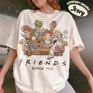 May include: An ivory short-sleeve t-shirt featuring a graphic print of Toy Story characters sitting on a couch. The text below the characters reads "FRIENDS SINCE 1995". A circular graphic in the top right corner contains the text "COMFORT COLORS IVORY".
