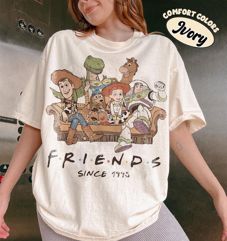 May include: An ivory short-sleeve t-shirt featuring a graphic print of Toy Story characters sitting on a couch. The text below the characters reads "FRIENDS SINCE 1995". A circular graphic in the top right corner contains the text "COMFORT COLORS IVORY".