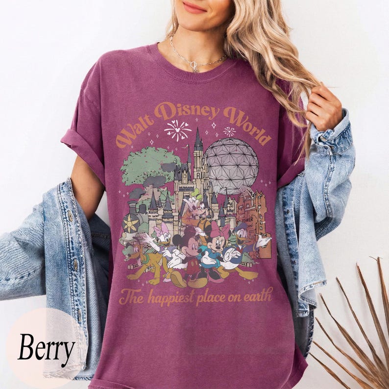 May include: Berry-colored t-shirt featuring a graphic of Walt Disney World characters and landmarks. The design includes Mickey Mouse, Donald Duck, and Cinderella's castle. Text reads "Walt Disney World" and "The happiest place on earth."