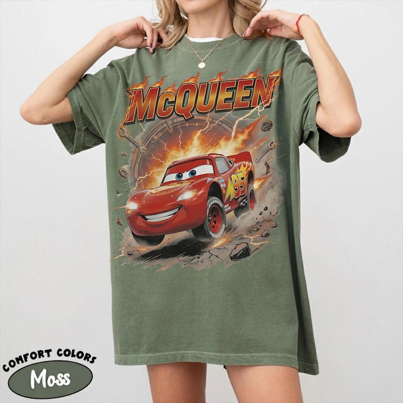 Może przedstawiać: Koszulka w kolorze mchu z grafiką Zygzaka McQueena z filmu Auta. Projekt zawiera napis "McQueen" w płomiennej czcionce i numer 95 na samochodzie. Koszulka jest marki Comfort Colors.
