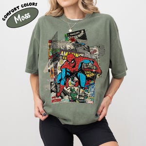 Pode incluir: Camiseta verde-oliva com um gr&aacute;fico do Homem-Aranha em um collage de capas de quadrinhos. As palavras "COMFORT COLORS" e "Moss" s&atilde;o impressas no canto superior esquerdo da camiseta. A camiseta tem um corte confort&aacute;vel.