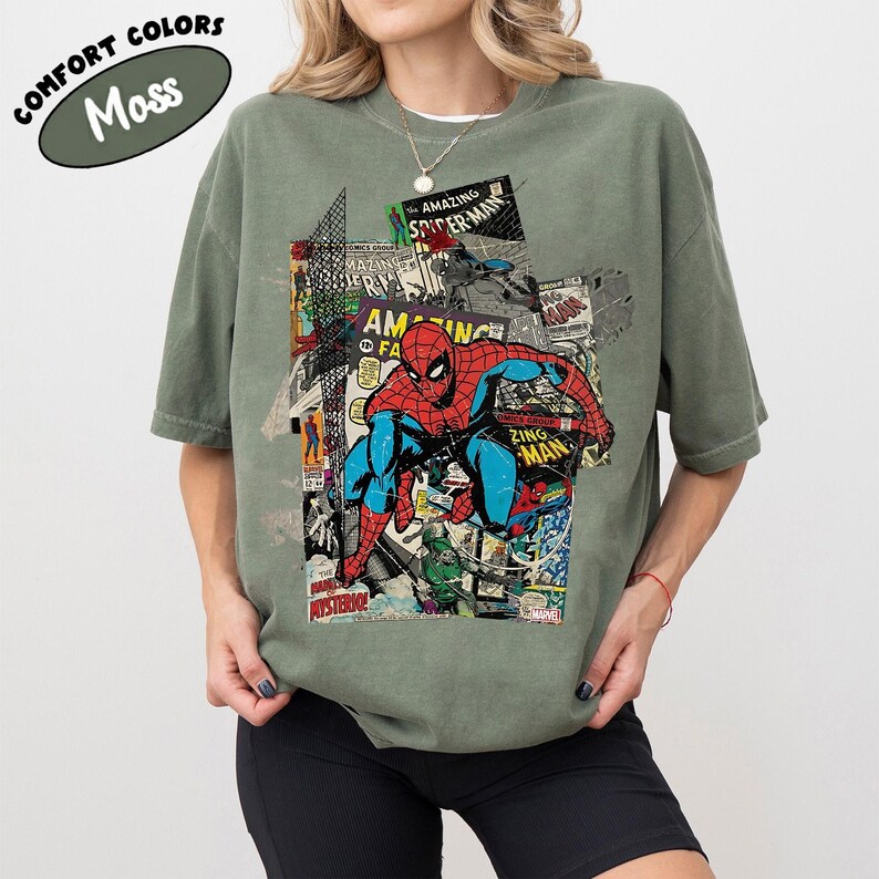Pode incluir: Camiseta verde-oliva com um gr&aacute;fico do Homem-Aranha em um collage de capas de quadrinhos. As palavras "COMFORT COLORS" e "Moss" s&atilde;o impressas no canto superior esquerdo da camiseta. A camiseta tem um corte confort&aacute;vel.