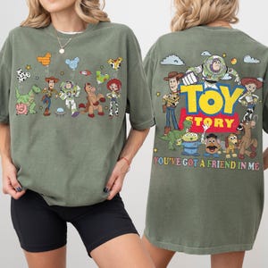 May include: An olive green t-shirt featuring Toy Story characters. The front displays Woody, Buzz Lightyear, Rex, Hamm, Bullseye, and Jessie with balloons. The back shows the Toy Story logo and characters with the text, You've Got A Friend In Me.