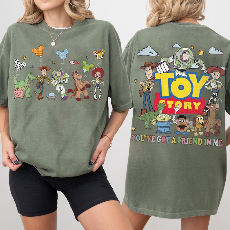 May include: An olive green t-shirt featuring Toy Story characters. The front displays Woody, Buzz Lightyear, Rex, Hamm, Bullseye, and Jessie with balloons. The back shows the Toy Story logo and characters with the text, You've Got A Friend In Me.