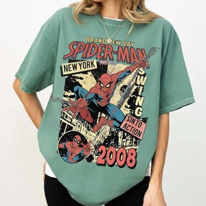 May include: Green t-shirt with a Spider-Man comic book design. The graphic includes the words "Brand New Day", "Spider-Man", "New York", and "Swing Into Action". The design also features the year "2008" and an illustration of Spider-Man.