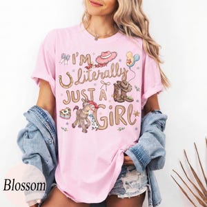 K&ouml;nnte beinhalten: Hellrosa T-Shirt mit dem Text "I'm Literally Just a Girl" und Cowboy-Grafiken. Das Design umfasst einen Cowboyhut, Stiefel, ein Pferd und Sterne. Das Shirt hat eine hellrosa Farbe.