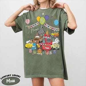K&ouml;nnte beinhalten: Moosgr&uuml;nes T-Shirt mit einer farbenfrohen Grafik von Zeichentrickautos, Luftballons und dem Text "HAPPY BIRTHDAY" mit einem "5" Luftballon. Das Shirt enth&auml;lt auch die Texte "Name" und "95". Das Shirt ist von der Marke Comfort Colors.