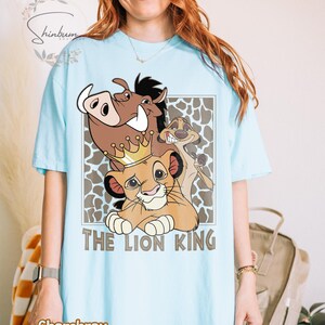 May include: Light blue t-shirt featuring a graphic of characters from The Lion King, including Simba wearing a crown, Pumbaa, and Timon. The words "THE LION KING" are printed below the characters. The shirt is made of chambray material.