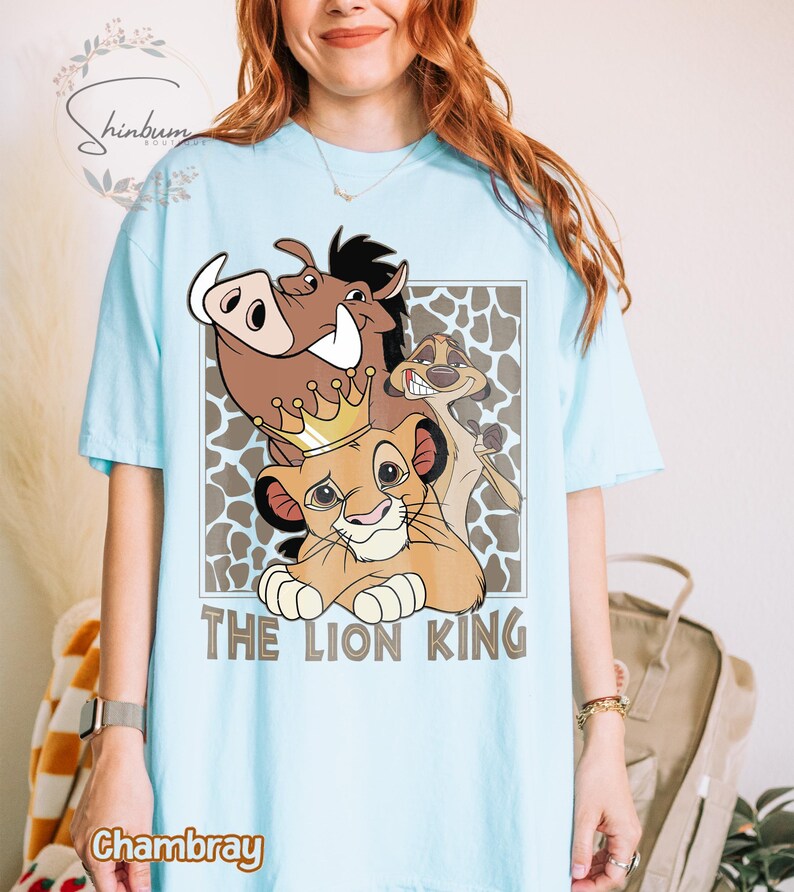 May include: Light blue t-shirt featuring a graphic of characters from The Lion King, including Simba wearing a crown, Pumbaa, and Timon. The words "THE LION KING" are printed below the characters. The shirt is made of chambray material.