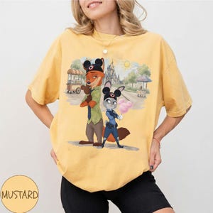 Puede incluir: Camiseta amarilla mostaza con una ilustraci&oacute;n de dibujos animados de Nick Wilde y Judy Hopps de Zootopia, con orejas de Mickey Mouse. Los personajes est&aacute;n frente a una escena de parque con un castillo y algod&oacute;n de az&uacute;car.