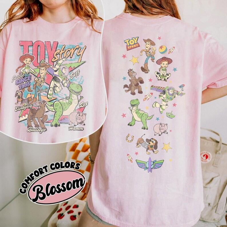 May include: Pink t-shirt featuring a colorful graphic of Toy Story characters, including Woody, Buzz Lightyear, and Rex. The front of the shirt displays the words "Toy Story" and "Jessie Since 1995". The back of the shirt has additional character graphics.