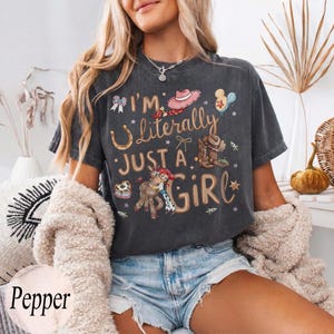 K&ouml;nnte beinhalten: Dunkelgraues T-Shirt mit dem Text "I'm Literally Just a Girl" und verschiedenen Cartoon-Grafiken, darunter ein Hufeisen, ein Cowboyhut und ein Pferd. Das Shirt wird mit Jeansshorts und einer cremefarbenen Strickjacke getragen.