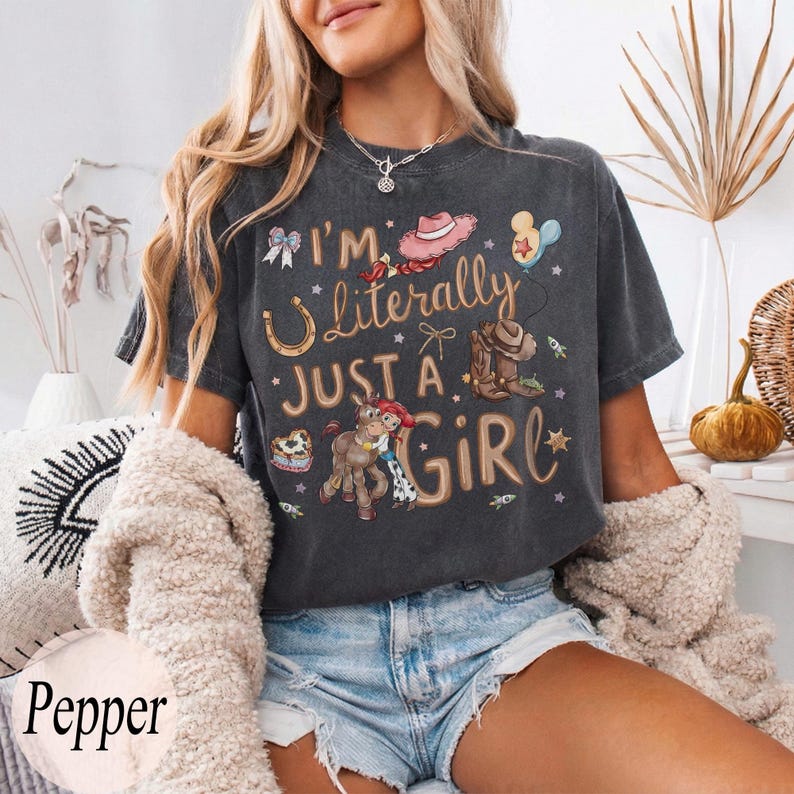 K&ouml;nnte beinhalten: Dunkelgraues T-Shirt mit dem Text "I'm Literally Just a Girl" und verschiedenen Cartoon-Grafiken, darunter ein Hufeisen, ein Cowboyhut und ein Pferd. Das Shirt wird mit Jeansshorts und einer cremefarbenen Strickjacke getragen.