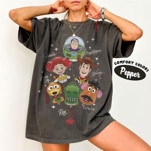 Puede incluir: Camiseta gris oscuro con un gr&aacute;fico de personajes de Toy Story, incluyendo a Buzz Lightyear, Woody, Jessie y el Sr. Potato. La camiseta tiene las palabras "Comfort Colors" y "Pepper" impresas en el lado derecho.