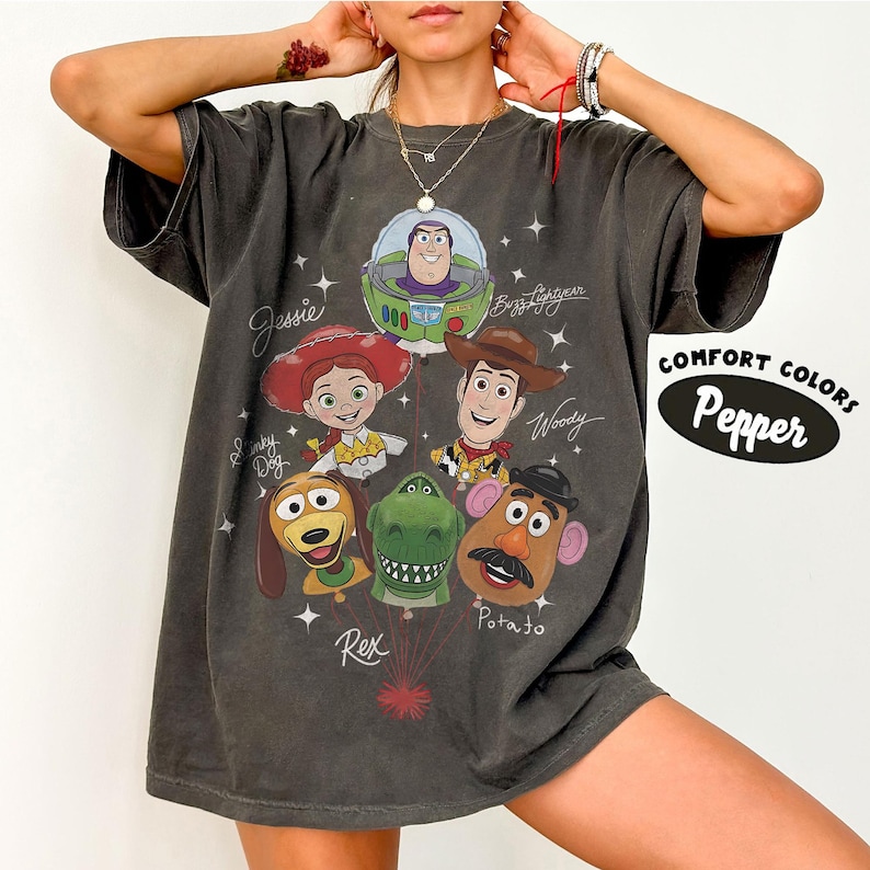 Puede incluir: Camiseta gris oscuro con un gr&aacute;fico de personajes de Toy Story, incluyendo a Buzz Lightyear, Woody, Jessie y el Sr. Potato. La camiseta tiene las palabras "Comfort Colors" y "Pepper" impresas en el lado derecho.