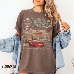 May include: Espresso-coloured t-shirt featuring a graphic of Radiator Springs with Lightning McQueen. The design includes the text "Radiator Springs" and "California est. 2012". The shirt has rolled-up sleeves.