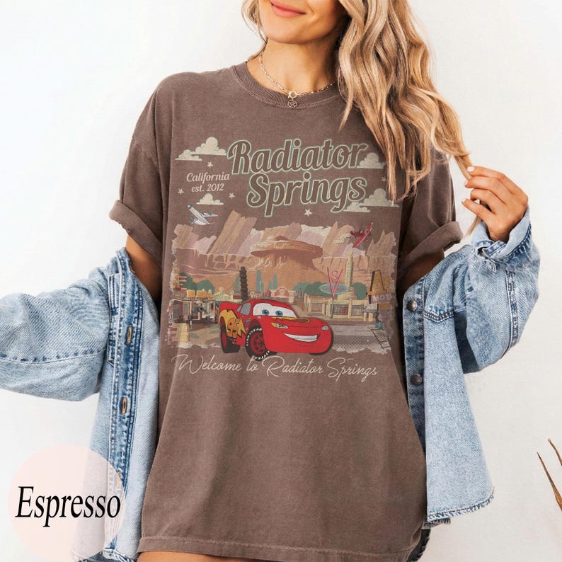 May include: Espresso-coloured t-shirt featuring a graphic of Radiator Springs with Lightning McQueen. The design includes the text "Radiator Springs" and "California est. 2012". The shirt has rolled-up sleeves.