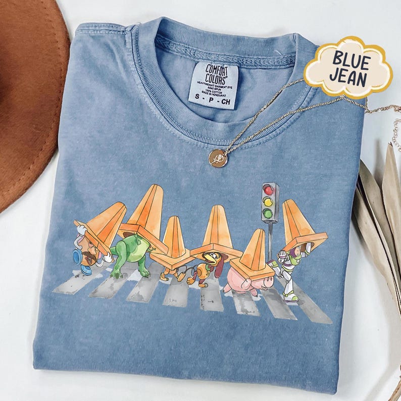May include: Blue jean colored t-shirt featuring a graphic of cartoon characters crossing a crosswalk, wearing orange traffic cones. The shirt has a Comfort Colors tag and a small gold necklace. The text "BLUE JEAN" is on a cloud-shaped tag.