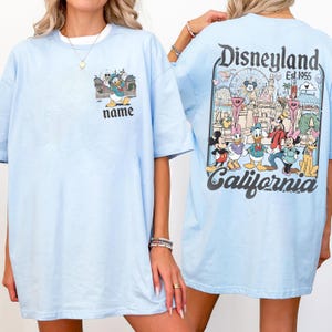 May include: Light blue t-shirt featuring a Disneyland design. The front of the shirt has a cartoon graphic of Donald Duck with the word "name" below. The back of the shirt displays a Disneyland graphic with various cartoon characters and the text "Disneyland Est. 1955 California."