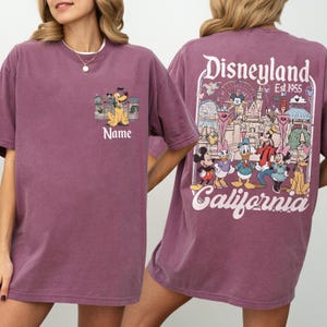 May include: A maroon t-shirt featuring a Disneyland design. The front pocket has a cartoon graphic with the word "Name" below it. The back of the shirt displays a Disneyland graphic with the text "Disneyland Est. 1955 California".