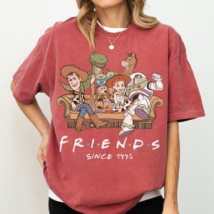 May include: A faded red short-sleeve t-shirt featuring a graphic print of Toy Story characters sitting on a couch. The text "FRIENDS" is printed in white with colorful dots between letters, followed by the text "SINCE 1995" in white below the couch.