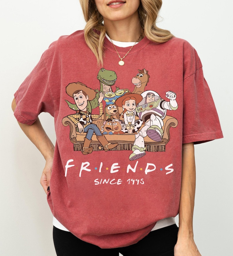 May include: A faded red short-sleeve t-shirt featuring a graphic print of Toy Story characters sitting on a couch. The text "FRIENDS" is printed in white with colorful dots between letters, followed by the text "SINCE 1995" in white below the couch.