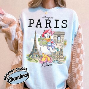 K&ouml;nnte beinhalten: Hellblaues Disneyland Paris T-Shirt mit Daisy Duck vor dem Eiffelturm und dem Arc de Triomphe. Das Shirt zeigt "Disneyland Paris" und "Paris". Die Worte "Comfort Colors Chambray" sind ebenfalls auf dem Shirt aufgedruckt.
