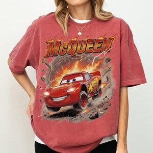 Może przedstawiać: Czerwona koszulka z grafiką Zygzaka McQueena z filmu Auta. Projekt zawiera słowo "McQueen" w kolorze pomarańczowym i ż&oacute;łtym, z samochodem wyścigowym i numerem 95. Koszulka jest w wyblakłym czerwonym kolorze.