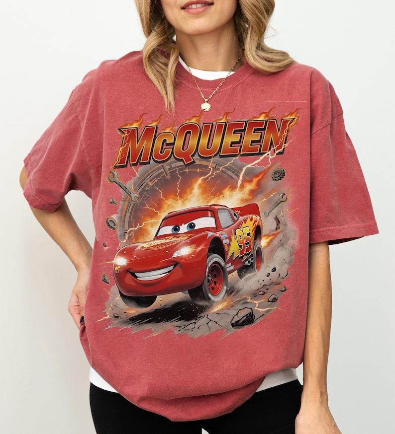 Może przedstawiać: Czerwona koszulka z grafiką Zygzaka McQueena z filmu Auta. Projekt zawiera słowo "McQueen" w kolorze pomarańczowym i ż&oacute;łtym, z samochodem wyścigowym i numerem 95. Koszulka jest w wyblakłym czerwonym kolorze.