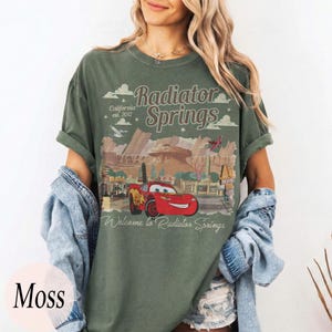 May include: Moss green t-shirt with a graphic of Radiator Springs from the film Cars. The design includes the text "Radiator Springs" and "Welcome to Radiator Springs" with a red car illustration. The shirt has a relaxed fit.
