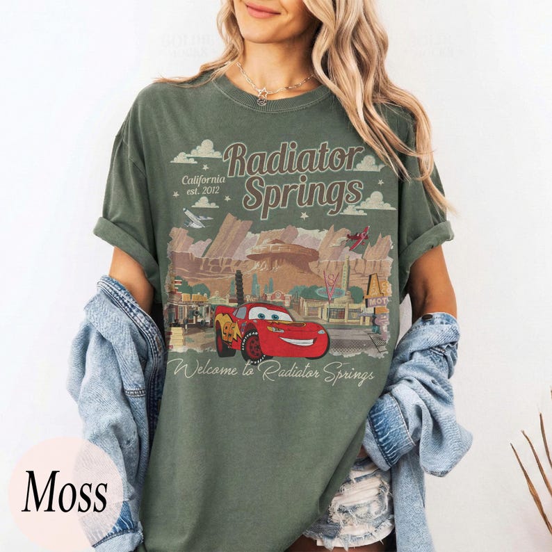 May include: Moss green t-shirt with a graphic of Radiator Springs from the film Cars. The design includes the text "Radiator Springs" and "Welcome to Radiator Springs" with a red car illustration. The shirt has a relaxed fit.