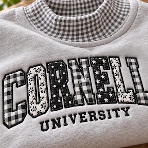 May include: A light gray sweatshirt with a mock neck collar featuring a black and white gingham pattern. The front displays the word CORNELL in large, appliqued letters with alternating gingham, polka dot, and star patterns, plus UNIVERSITY below.