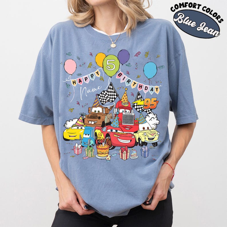 K&ouml;nnte beinhalten: Blaues Jeans-T-Shirt mit einem farbenfrohen Aufdruck von animierten Autos, die einen Geburtstag feiern. Das Design umfasst Luftballons, eine Geburtstagstorte und den Text "Happy Birthday" und "Name". Das Shirt hat auch den Text "Comfort Colors".