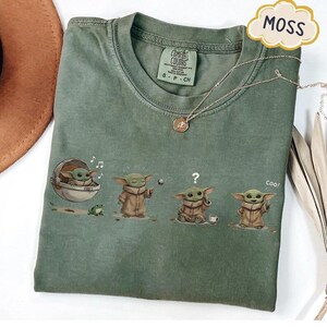 Puede incluir: Camiseta Comfort Colors verde musgo con un gr&aacute;fico de un personaje de una popular serie de ciencia ficci&oacute;n. El dise&ntilde;o muestra al personaje en diferentes poses. Tambi&eacute;n se ve un collar plateado con un colgante.
