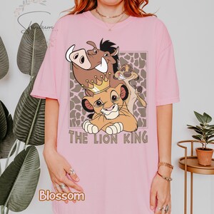 May include: Pink t-shirt featuring a graphic of characters from The Lion King, including Simba wearing a crown, Pumbaa, and Timon. The text "THE LION KING" is below the characters. The shirt has short sleeves.