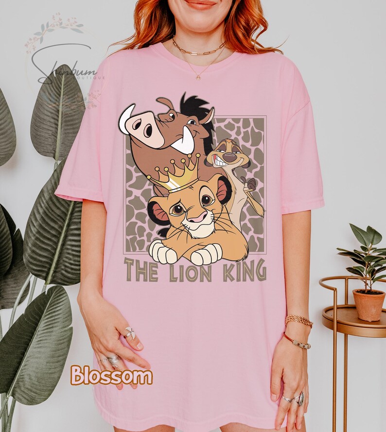 May include: Pink t-shirt featuring a graphic of characters from The Lion King, including Simba wearing a crown, Pumbaa, and Timon. The text "THE LION KING" is below the characters. The shirt has short sleeves.
