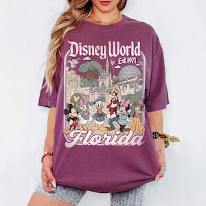 May include: A purple t-shirt featuring a graphic of Disney World in Florida. The design includes Mickey Mouse, Donald Duck, Goofy, and other characters, with the words "Disney World Est. 1971" and "Florida" in a vintage style.
