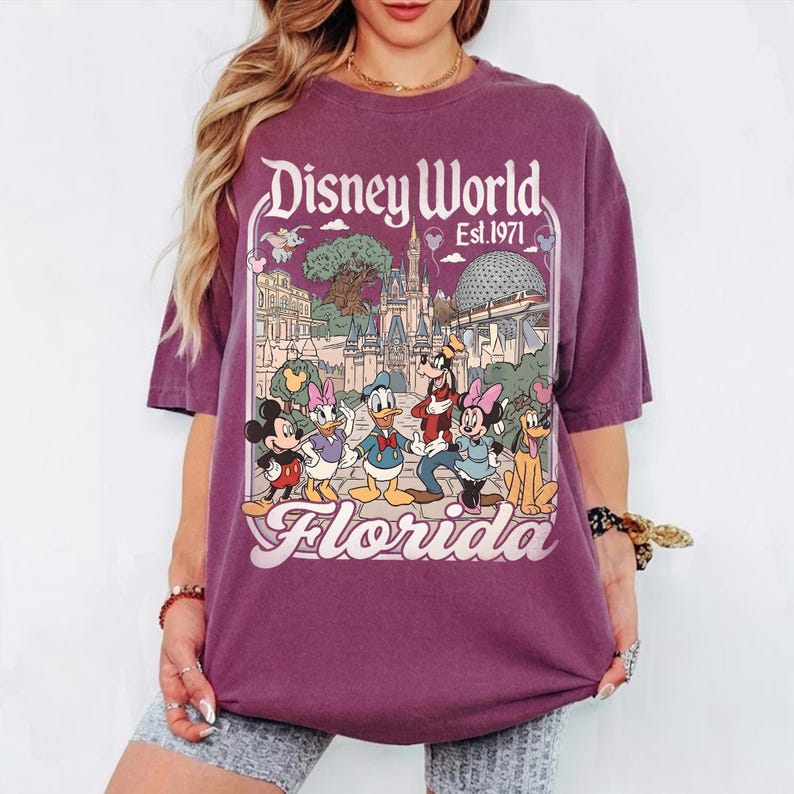 May include: A purple t-shirt featuring a graphic of Disney World in Florida. The design includes Mickey Mouse, Donald Duck, Goofy, and other characters, with the words "Disney World Est. 1971" and "Florida" in a vintage style.