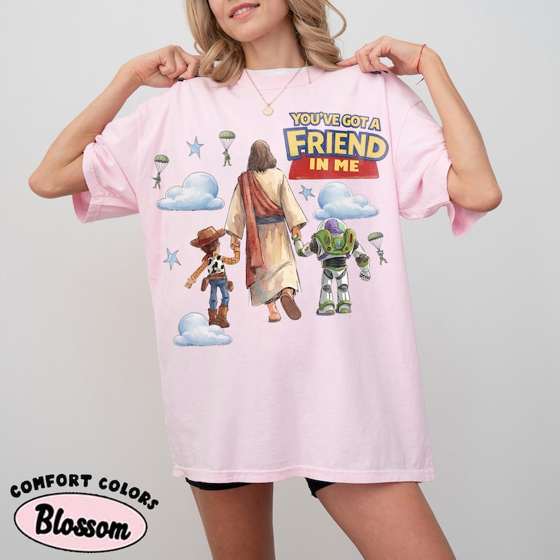 May include: A pale pink t-shirt featuring the phrase "YOU'VE GOT A FRIEND IN ME" and a graphic of Jesus, Woody, and Buzz Lightyear. The design includes clouds, stars, and parachutes. The shirt is labelled "Comfort Colors Blossom".