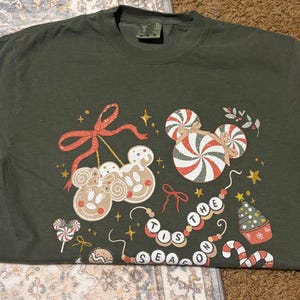 May include: Dark green t-shirt featuring a festive design with gingerbread Mickey Mouse heads, candy canes, and the phrase "THE TIS THE SEASON". The design includes red bows, stars, and a Christmas tree. A fun holiday themed tee.