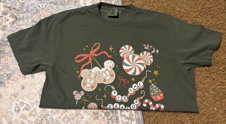 May include: Dark green t-shirt featuring a festive design with gingerbread Mickey Mouse heads, candy canes, and the phrase "THE TIS THE SEASON". The design includes red bows, stars, and a Christmas tree. A fun holiday themed tee.