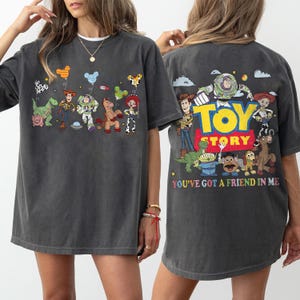 May include: A charcoal gray t-shirt featuring Toy Story characters. The front displays Woody, Buzz Lightyear, Rex, Hamm, Bullseye, and Jessie with Mickey-shaped balloons. The back shows the Toy Story logo and text reading YOU'VE GOT A FRIEND IN ME.