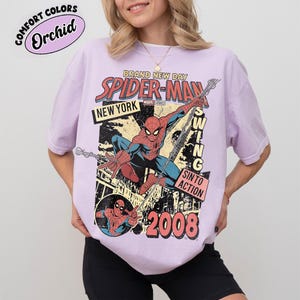 May include: A lilac t-shirt with a Spider-Man graphic, including the text "BRAND NEW DAY SPIDER-MAN NEW YORK" and the year "2008". The design also features "SWING INTO ACTION". The shirt is labeled "Comfort Colors Orchid".