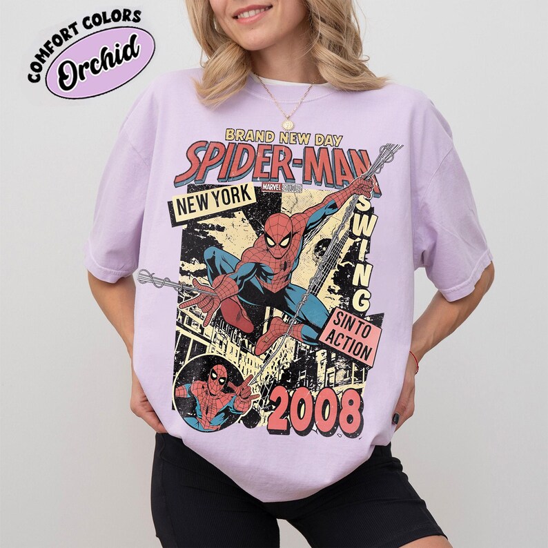 May include: A lilac t-shirt with a Spider-Man graphic, including the text "BRAND NEW DAY SPIDER-MAN NEW YORK" and the year "2008". The design also features "SWING INTO ACTION". The shirt is labeled "Comfort Colors Orchid".