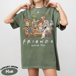 May include: A moss green short-sleeve t-shirt featuring a graphic of Toy Story characters sitting on a couch with the text "FRIENDS SINCE 1995" printed below. The characters include Woody, Buzz Lightyear, Jessie, Rex, Bullseye, and others.